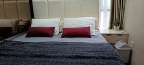 A bed with a headboard, two white pillows, two red pillows, a dark blue blanket, a bedside table with a remote control, a potted plant, and a trash bin. - Guesthouse Puri at Kemang (Jakarta Selatan)