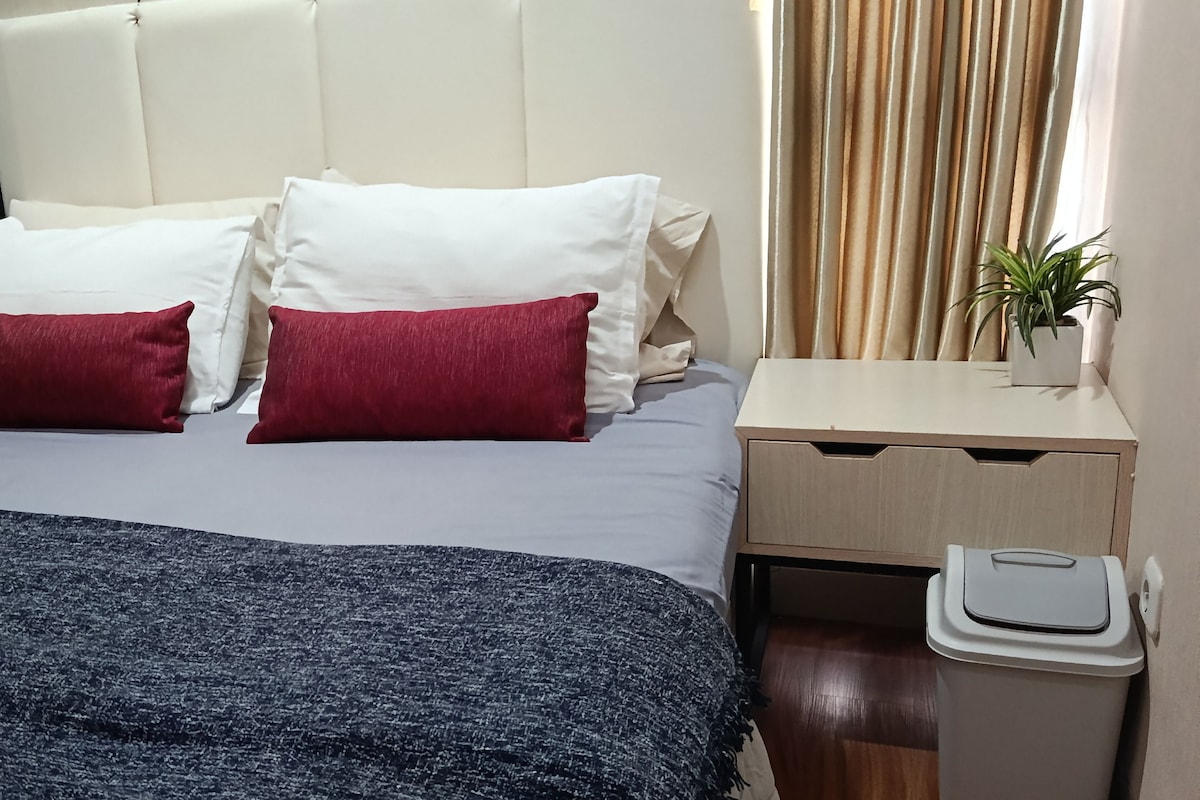A hotel room with a bed, pillows, a bedside table, a trash bin, and a potted plant.