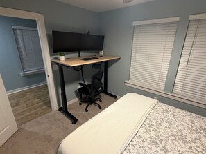 A bedroom with a bed, a desk with a computer, a chair, and a window with blinds. - Cane Bay / Summerville getaway—where Southern hospitality meets modern comfort. (Summerville)