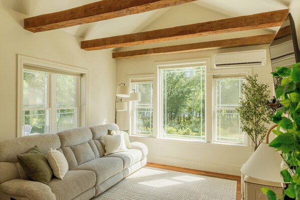 A room with a sofa, large windows, wooden beams, and an air conditioning unit.