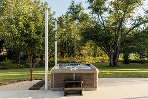 Una vasca idromassaggio con rivestimento in pietra, circondata da un solarium in cemento, immersa in un giardino curato con alberi e cielo sereno.
