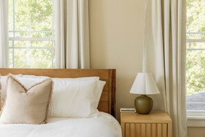 Ein Bett mit weißer Bettwäsche und zwei Kissen, ein Holz-Kopfteil, ein Nachttisch mit Lampe und ein Fenster mit Vorhängen.