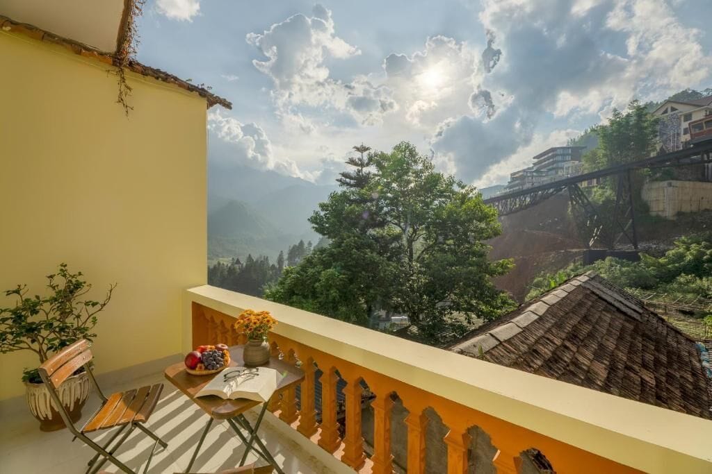 A balcony with a view of a bridge and mountains, a table with a bowl of fruit, and a potted plant.