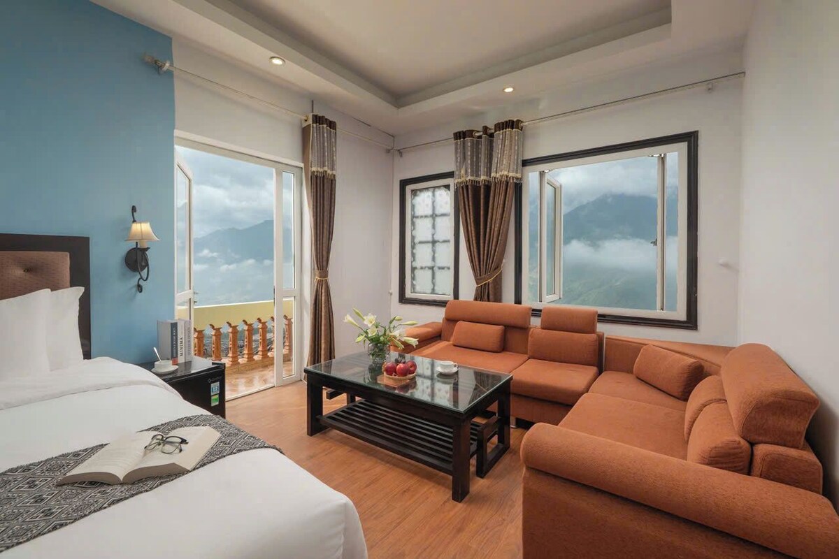 A hotel room with a bed, a sofa, a coffee table, and a view of mountains through large windows.