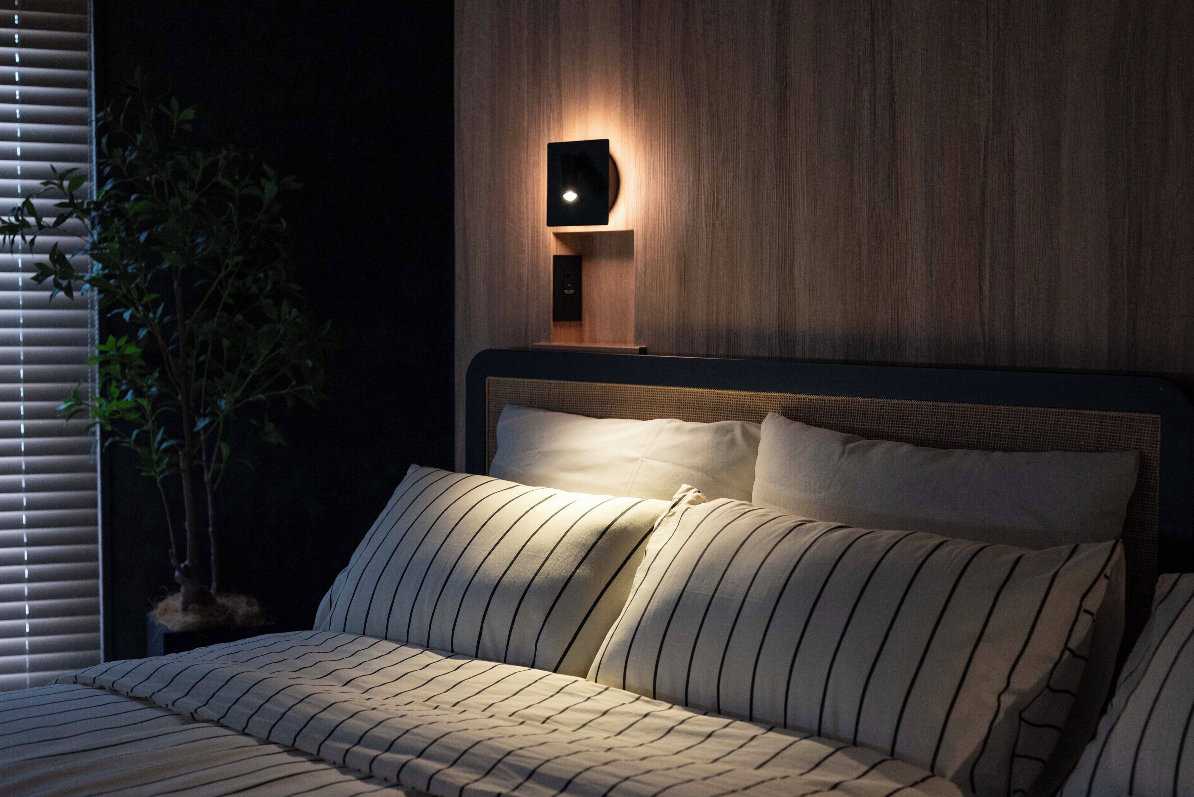 A bed with striped bedding, a wall-mounted light, and a potted plant.
