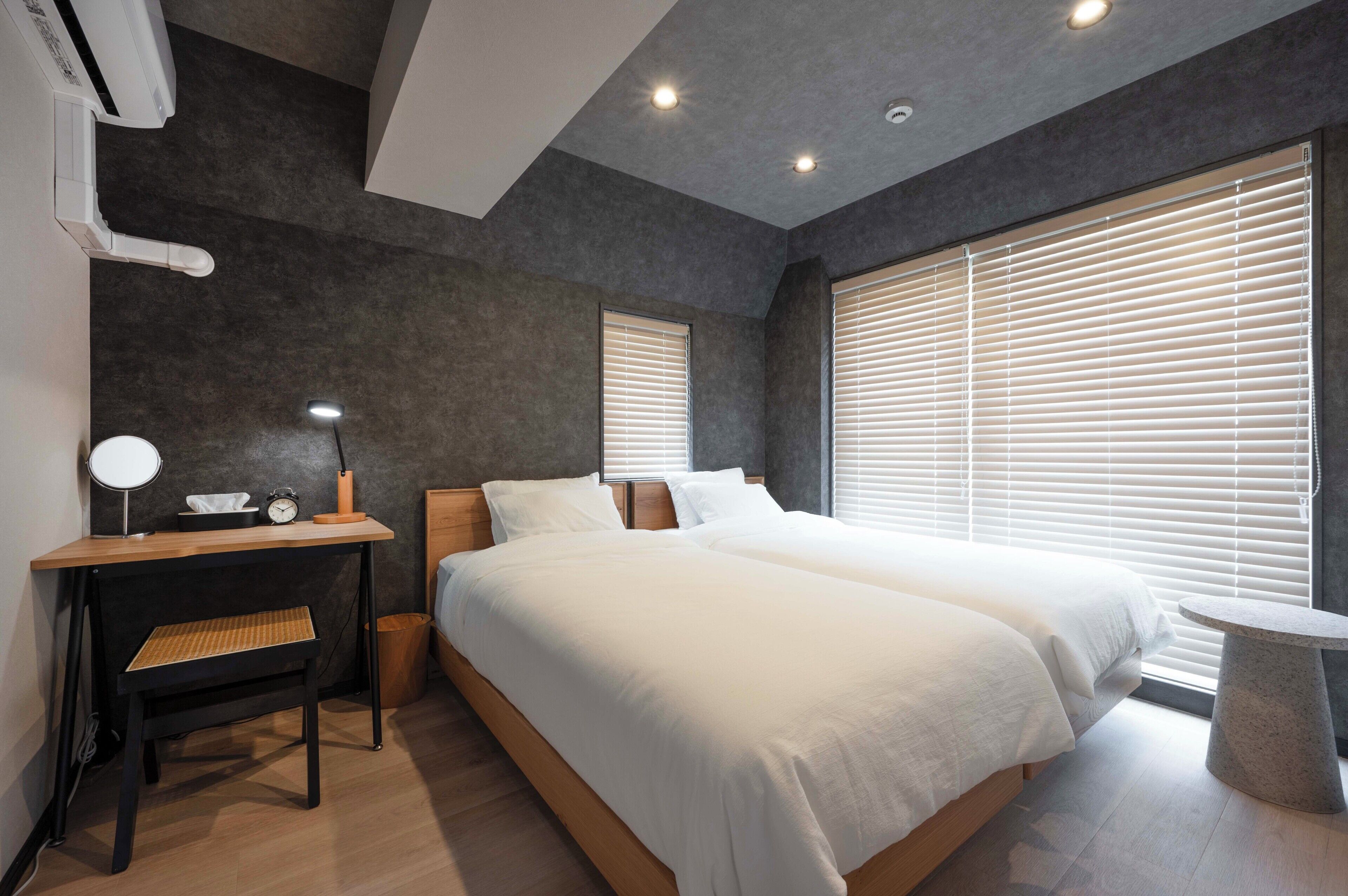 A modern bedroom with a large bed, bedside tables, a desk with a lamp, and a window with blinds.