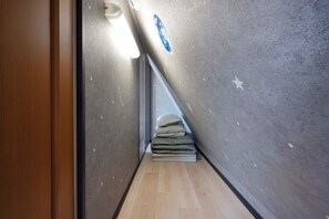 Um corredor estreito com teto inclinado, uma porta de madeira à esquerda e uma pilha de almofadas no final.