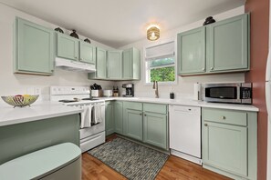 Una cocina con armarios verde claro, encimera blanca, estufa, microondas y ventana con vistas a la vegetación.