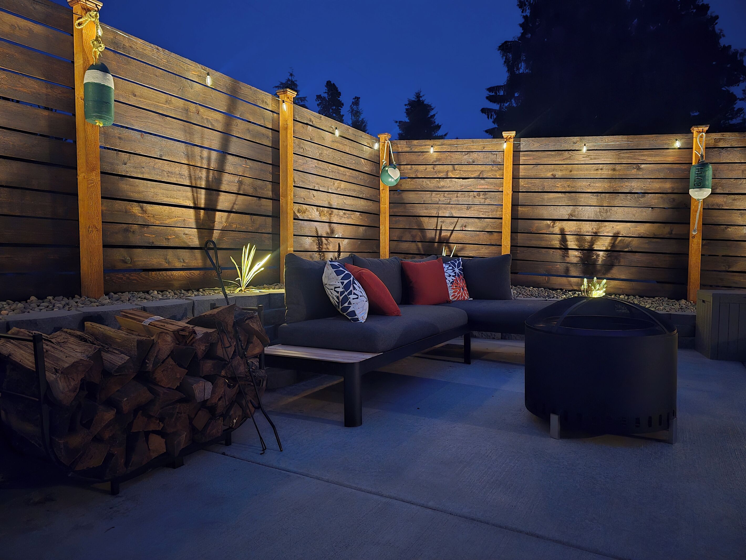 An outdoor seating area with a sofa and a fire pit, surrounded by a wooden fence and string lights.