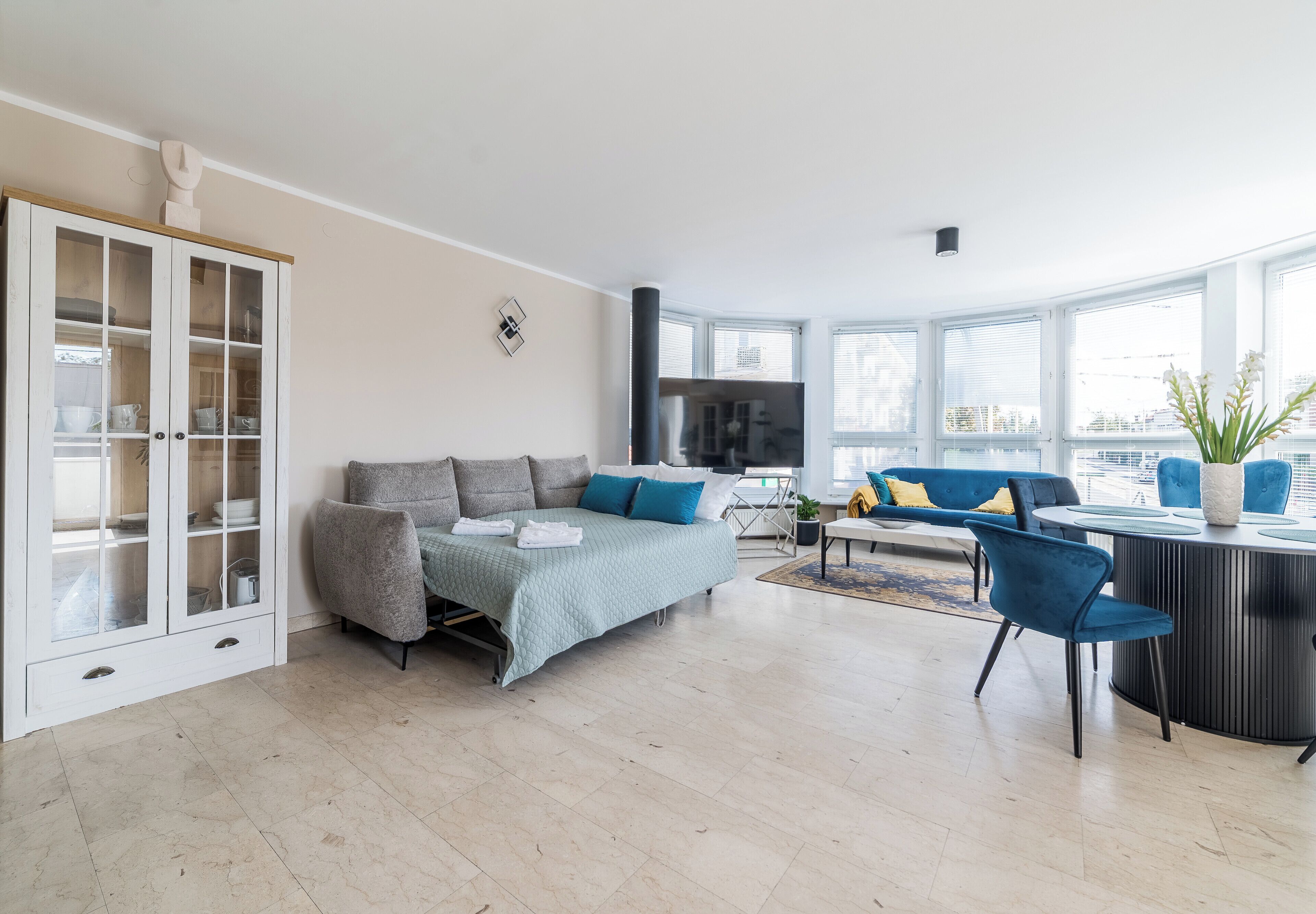 A modern living room with a sofa, a dining table with chairs, a cabinet with glass doors, and a large window providing a view of the city.