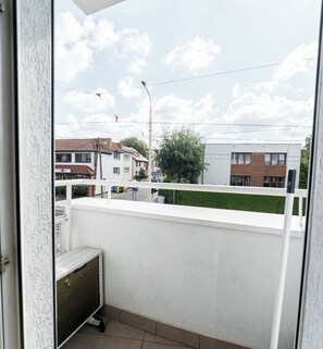 Ein Balkon mit weißer Reling, einem kleinen Tisch und Blick auf eine Straße mit Gebäuden und einem Baum.