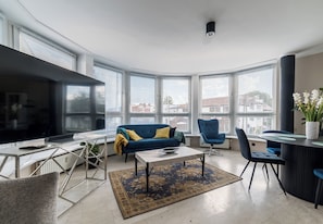 Ein modernes Wohnzimmer mit einem blauen Sofa, einem Couchtisch und einem Fernseher. Große Fenster mit Jalousien, eine Pflanze und ein Blick auf Gebäude im Freien.