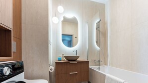 A modern bathroom with a built-in washing machine, a wooden vanity, a large mirror, and a bathtub. - Ogrody Tesoro 52 Apartment (Pogórze)