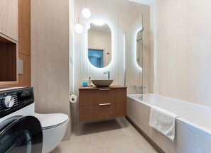 A modern bathroom with a built-in washing machine, a wooden vanity, a large mirror, and a bathtub. - Ogrody Tesoro 52 Apartment (Pogórze)