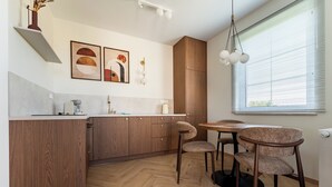 A modern kitchen with wooden cabinets, a dining area with a round table and chairs, and a window with blinds. - Ogrody Tesoro 52 Apartment (Pogórze)