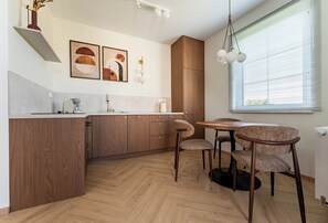 A modern kitchen with wooden cabinets, a dining area with a round table and chairs, and a window with blinds. - Ogrody Tesoro 52 Apartment (Pogórze)