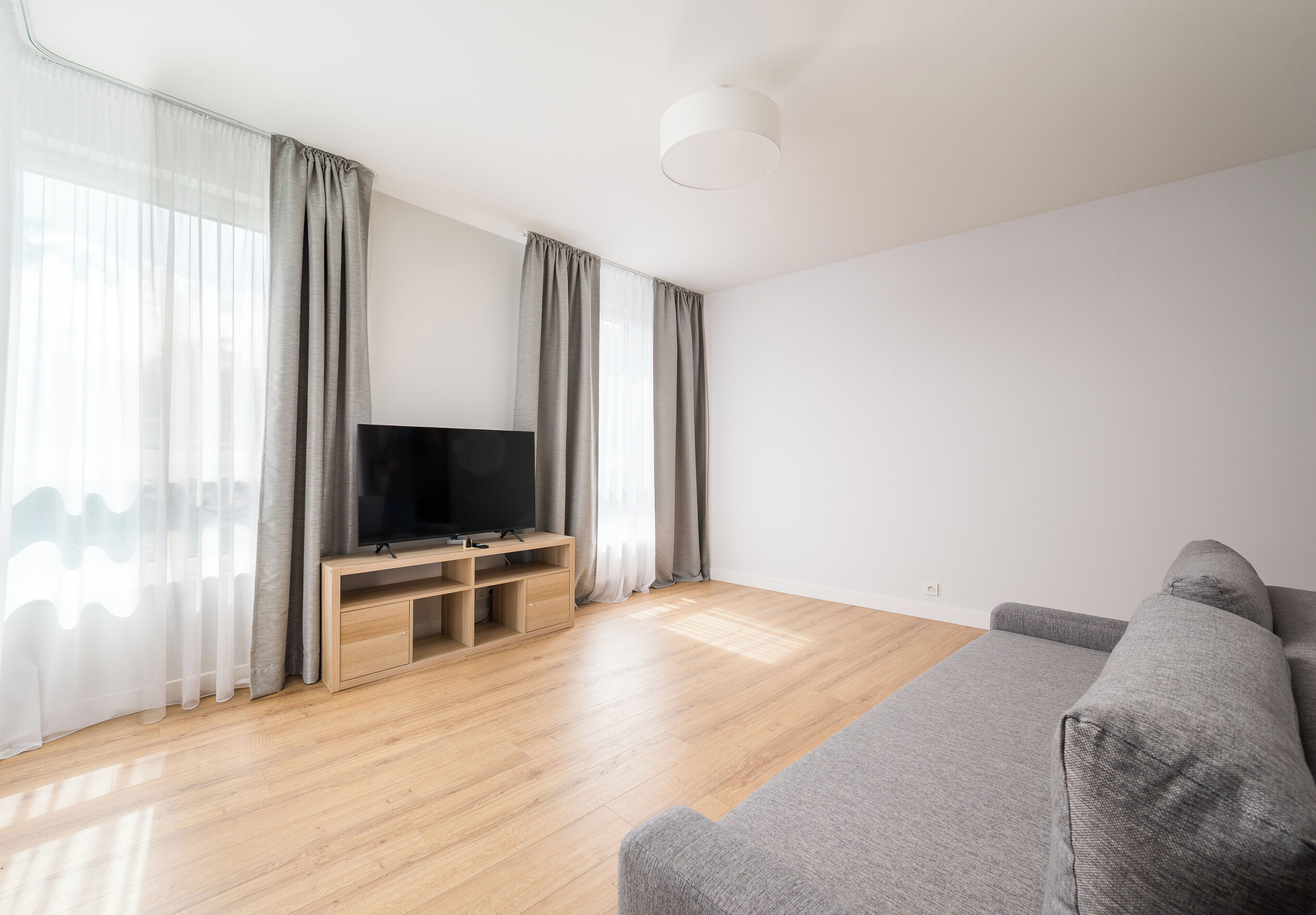 A modern living room with a flat-screen TV on a wooden stand, a grey sofa, and sheer curtains on the windows.