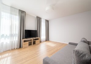 Ein modernes Wohnzimmer mit einem Flachbildfernseher auf einem Holzboden, einem grauen Sofa und leichten Vorhängen an den Fenstern.