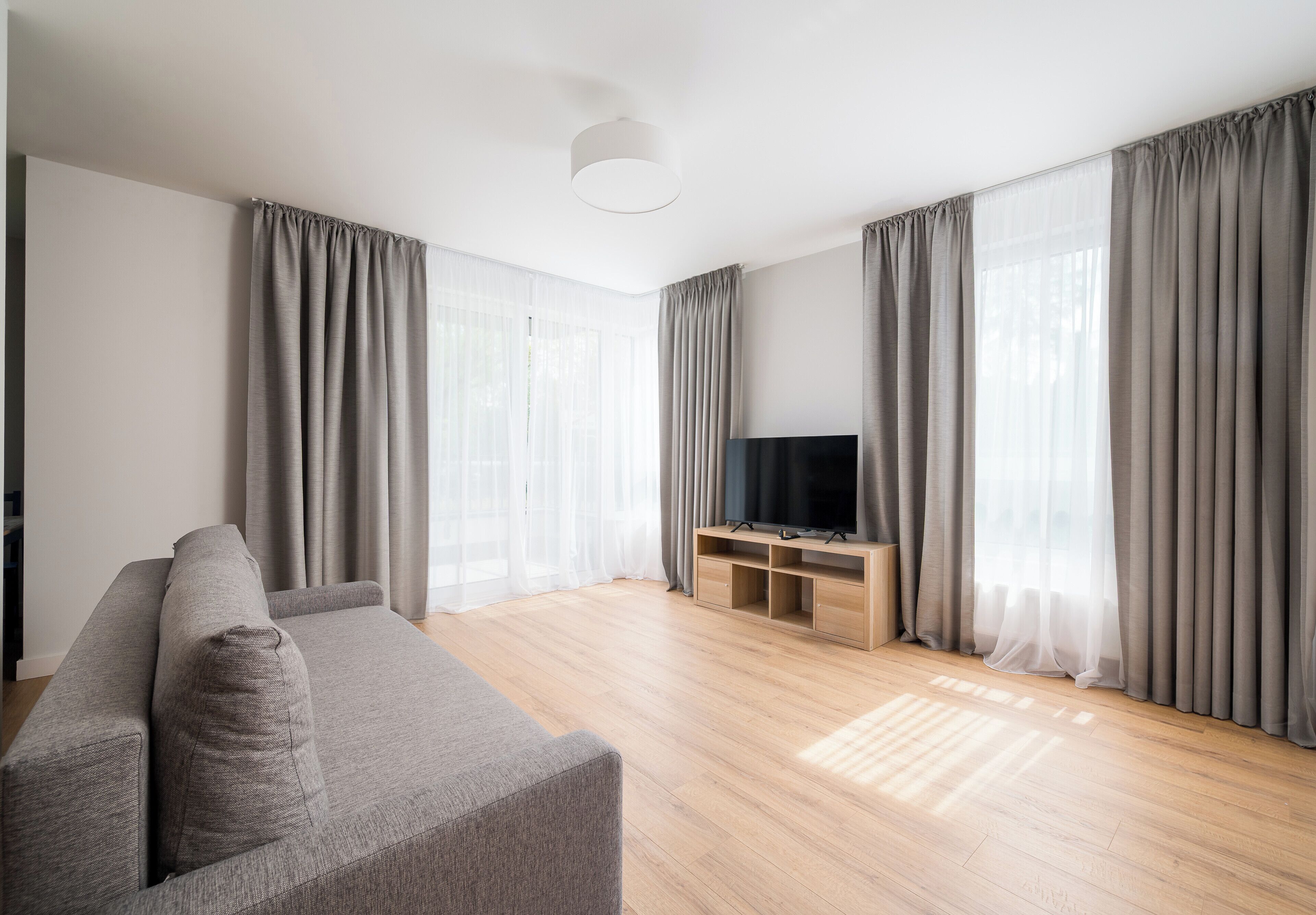En moderne stue med en grå sofa, et fjernsyn på et træstel og store vinduer med grå gardiner.