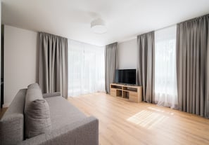 En moderne stue med en grå sofa, et fjernsyn på et træstel og store vinduer med grå gardiner.