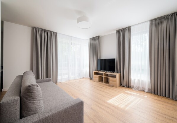 A modern living room with a grey sofa, a TV on a wooden stand, and large windows with grey curtains. - Warszawa Zacisze-Wilno Deluxe Apartment (Warszawa)