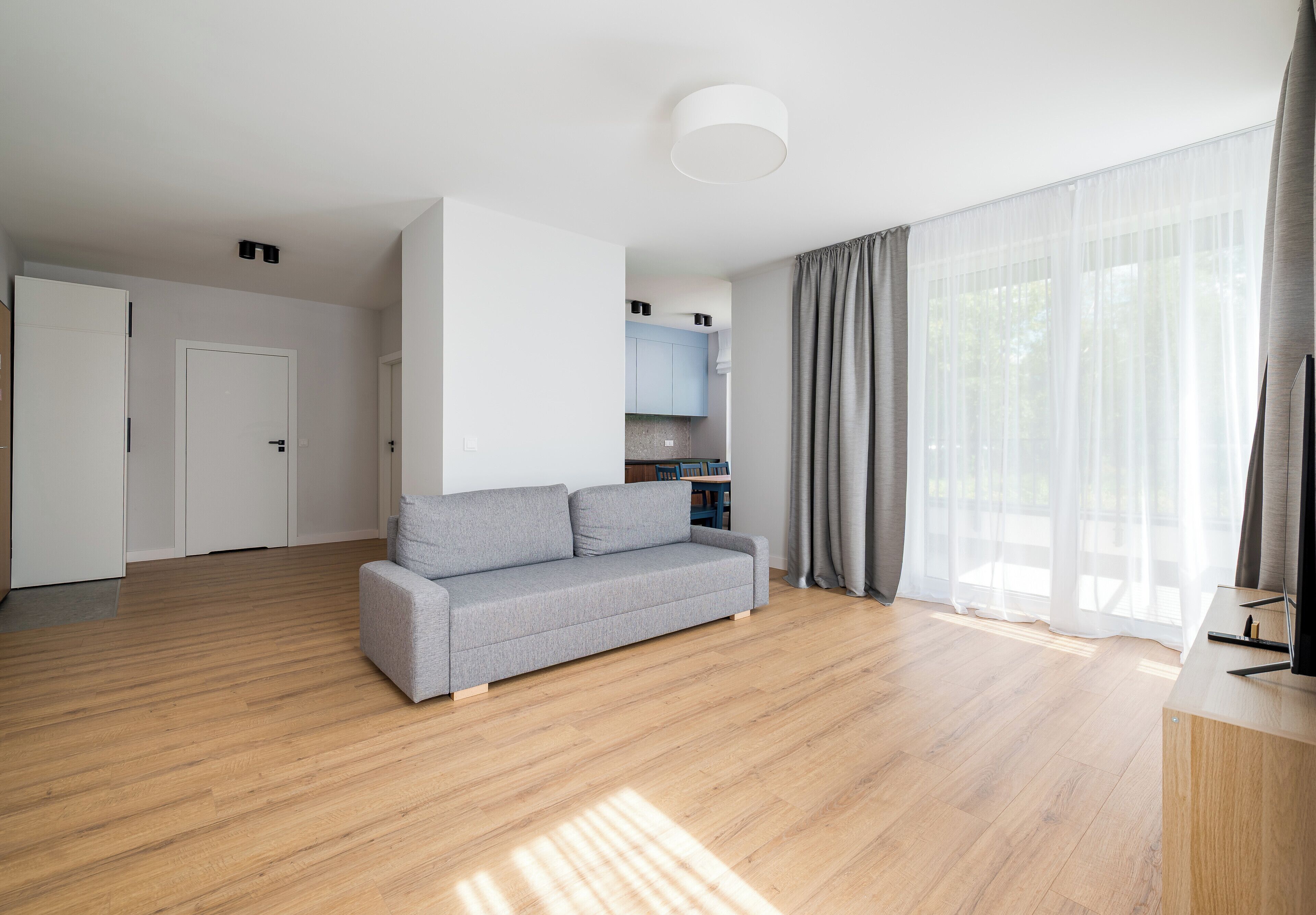 Ein geräumiges Wohnzimmer mit grauem Sofa, Holzböden, einem Essbereich mit Tisch und Stühlen sowie einer Küche mit blauen Schränken.