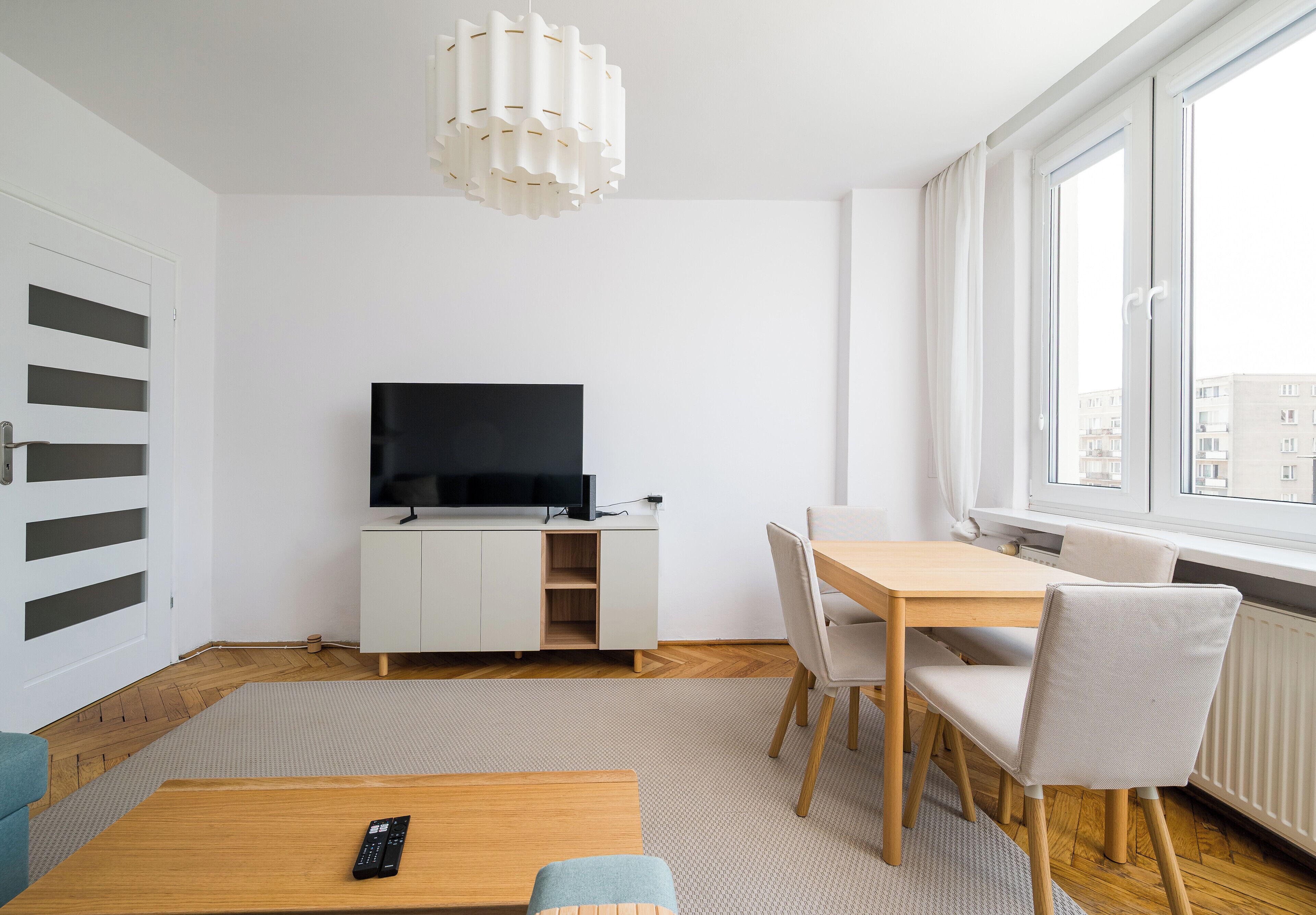 Ein modernes Wohnzimmer mit einem Flachbildfernseher auf einem weißen Schrank, einem Holzespeisetisch mit Stühlen und einem großen Fenster mit Blick auf Gebäude.