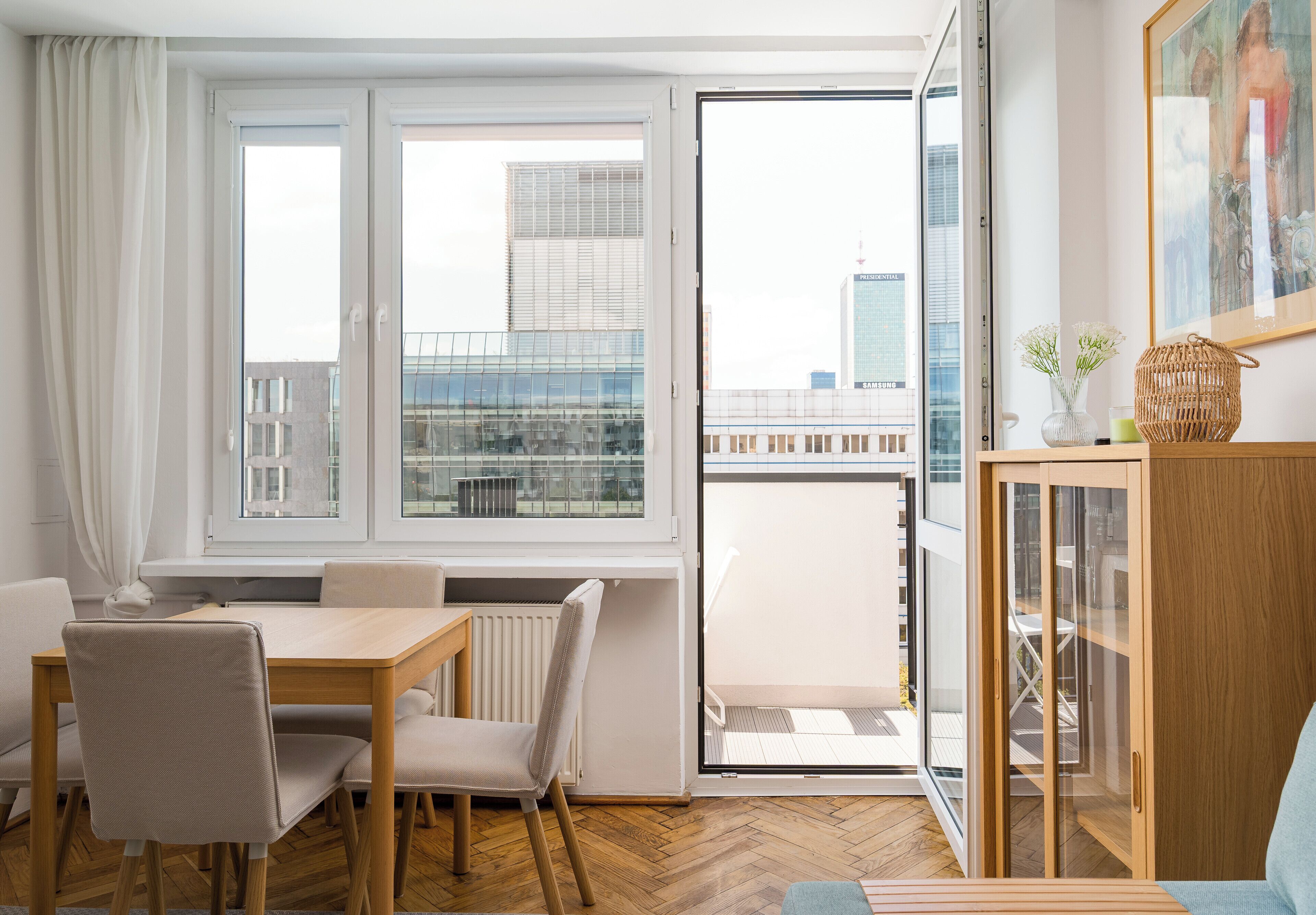 Een moderne woonkamer met een eethoek, een glazen deur naar een balkon en uitzicht op stadsgebouwen.