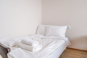 A single bed with white linens and two rolled towels, a white wall, and a wooden floor. - Park Kozielski Comfort Apartment (Gliwice)