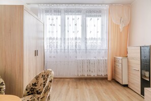 Una habitación con un armario de madera, una silla con estampado floral, una mesita pequeña y un mueble con cajones.