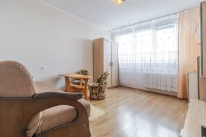 A living room with a sofa, a small table, a patterned armchair, a wardrobe, and a window with curtains. - Park Kozielski Comfort Apartment (Gliwice)