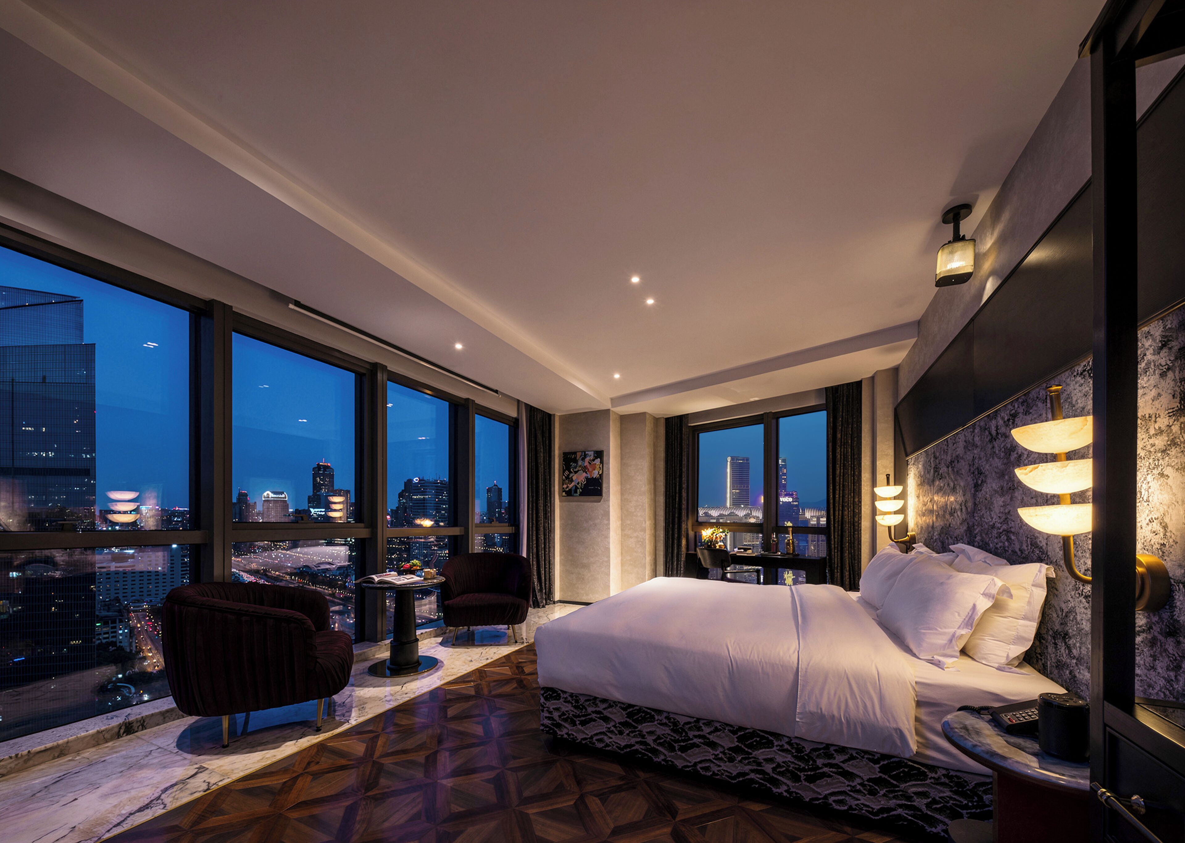 Habitación de hotel con una cama grande, un escritorio, una silla y vistas a la ciudad a través de amplias ventanas.