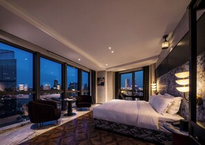 Une chambre d’hôtel avec un grand lit, un bureau, une chaise et une vue sur le paysage urbain grâce à de grandes fenêtres.