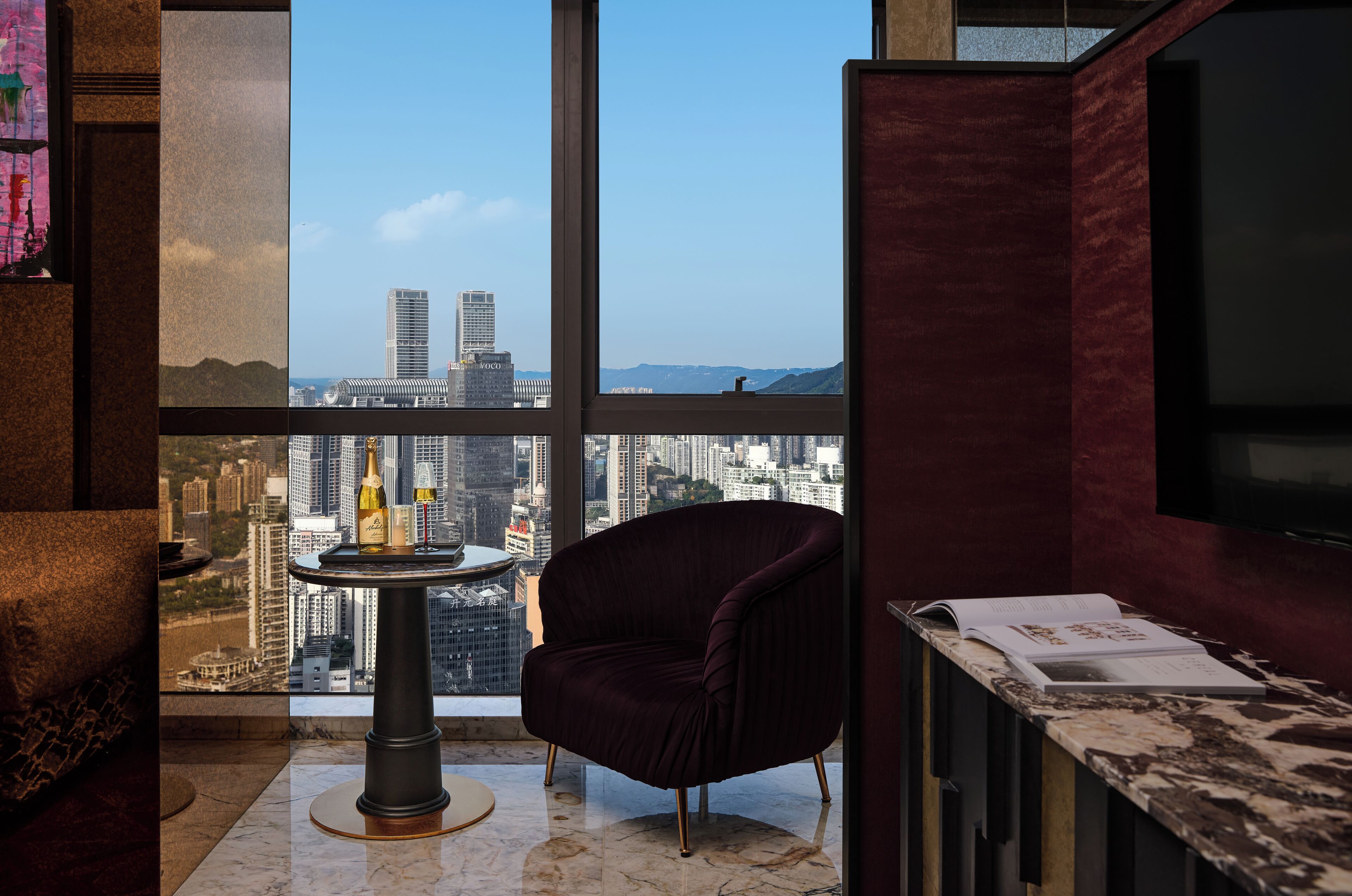 A room with a large window offering a city view, a round table with a bottle and glass, a dark armchair, and a marble floor.