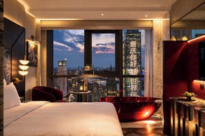 Une chambre d’hôtel moderne avec un grand lit, un fauteuil rouge, une petite table avec une bouteille et une bougie, et une vue sur la ville à travers de grandes fenêtres du sol au plafond.