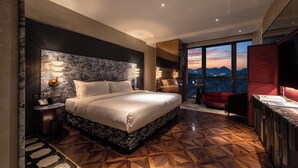 Une chambre d’hôtel moderne avec un grand lit, un coin salon avec un fauteuil rouge et une vue sur les montagnes par la fenêtre.