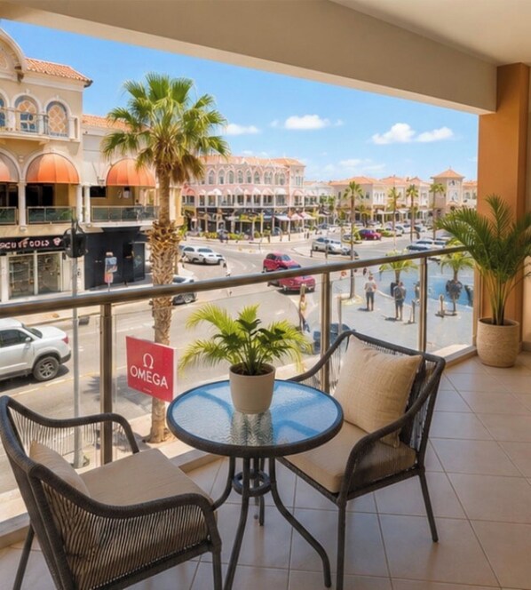 A balcony with a round table and chairs, overlooking a street with palm trees and a sign for 'OMEGA.' - Enjoyable 1-bedroom apartment with AC in lovely Oranjestad (Oranjestad)