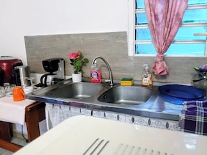 A kitchen with a double sink, a coffee maker, a potted plant, and a window with a curtain. - LE MILANO (Kourou)