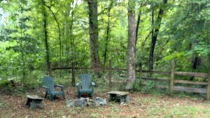 A forested area with outdoor seating including two Adirondack chairs and a small table.