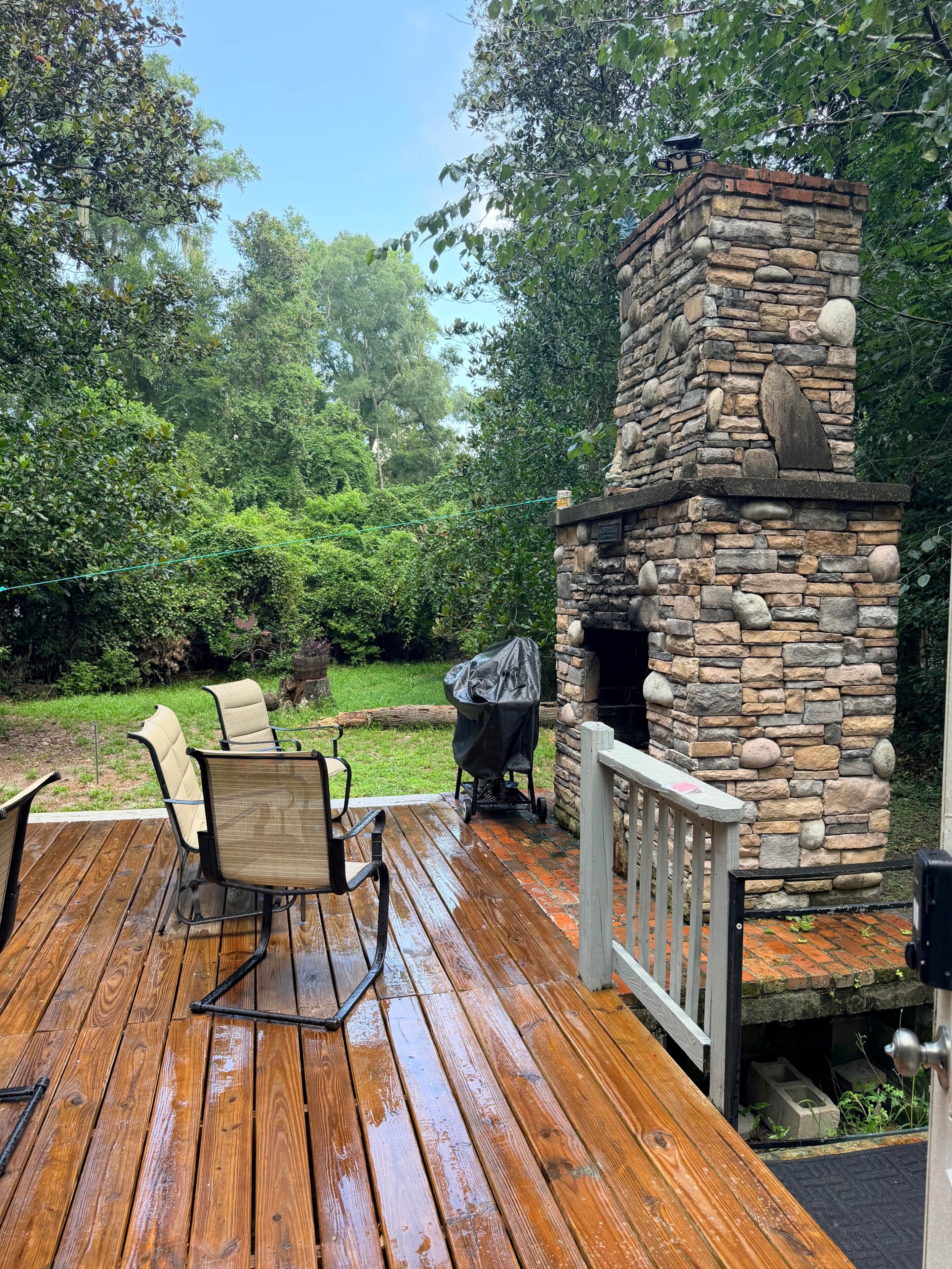 Una terraza de madera con chimenea de piedra, mobiliario exterior y una zona de parrilas techada.