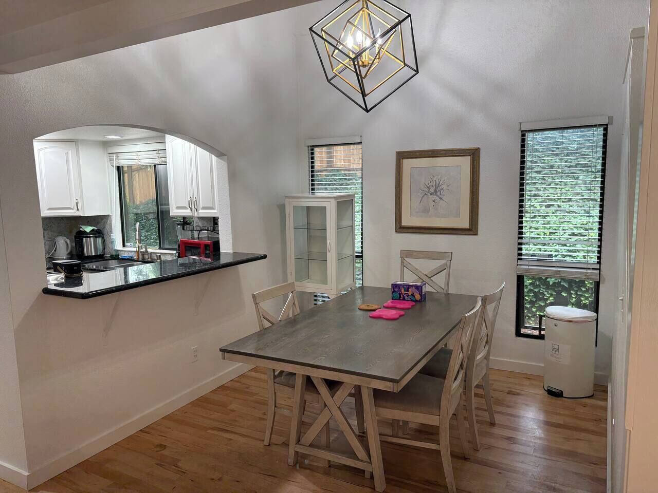 A dining area with a table and chairs, a kitchen with white cabinets, and a window with blinds.