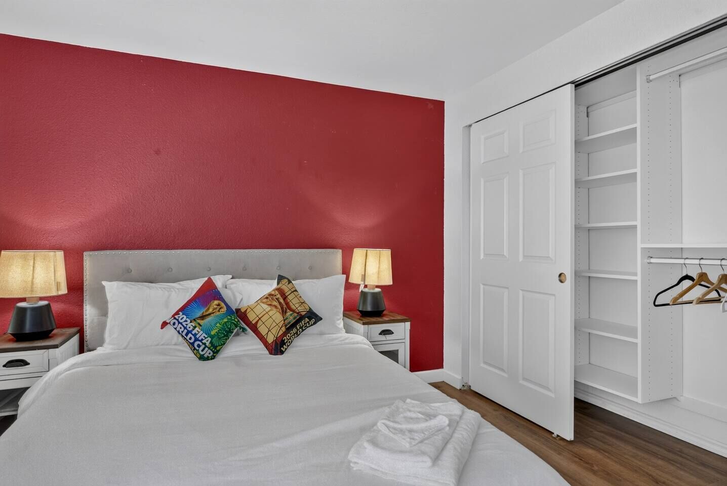 A hotel room with a red wall, a white bed, two nightstands with lamps, and a white closet with hangers.