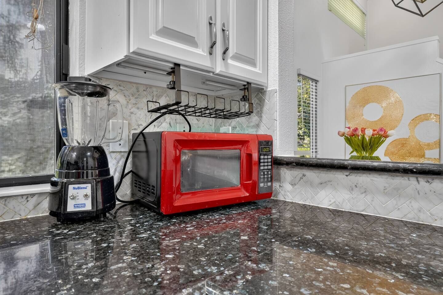 A kitchen with a red microwave, a blender, and a window with a view of trees.
