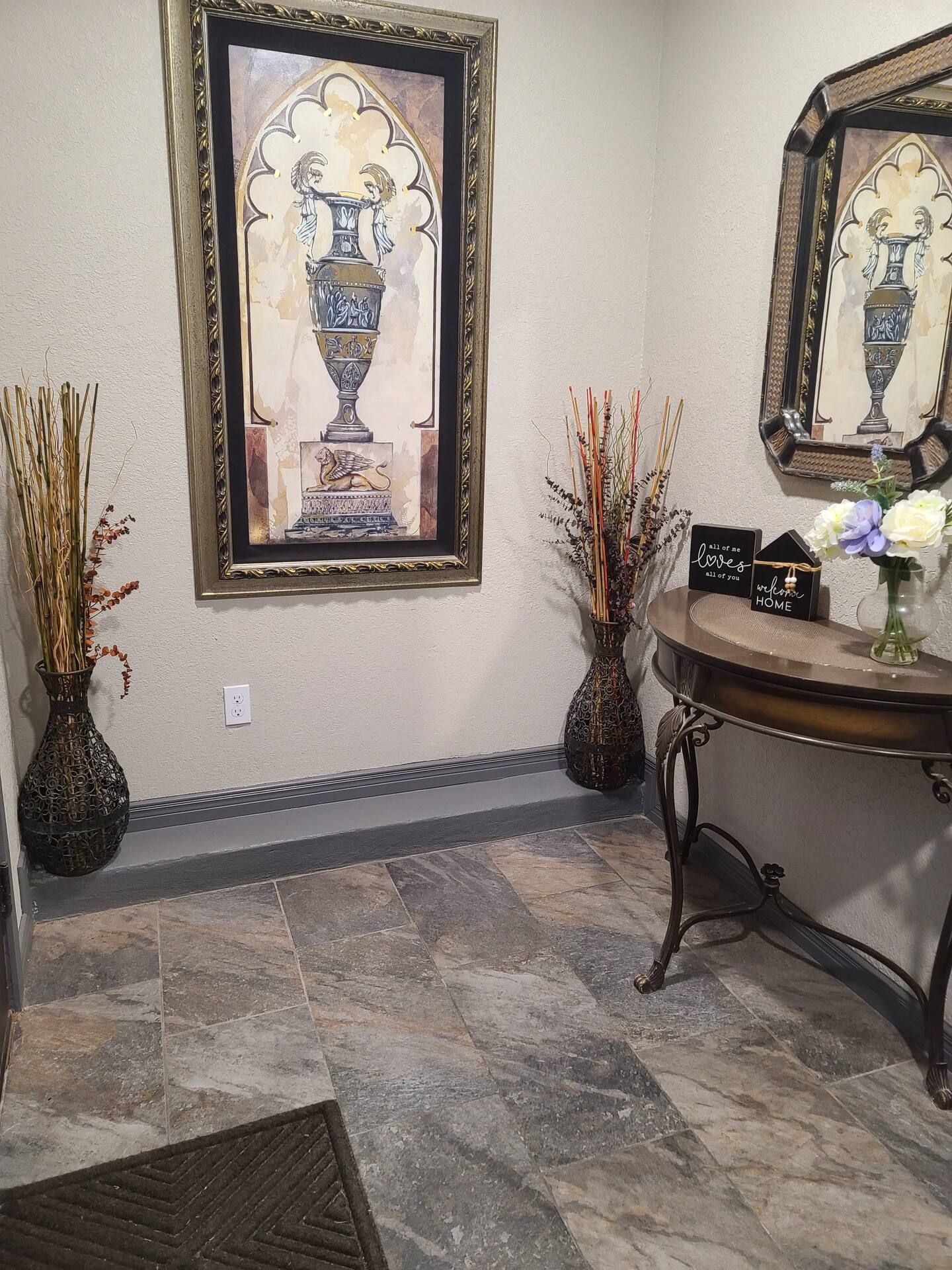 A hallway with a framed artwork of a vase, two vases with dried plants, a small table with a sign, and a mirror reflecting the artwork.