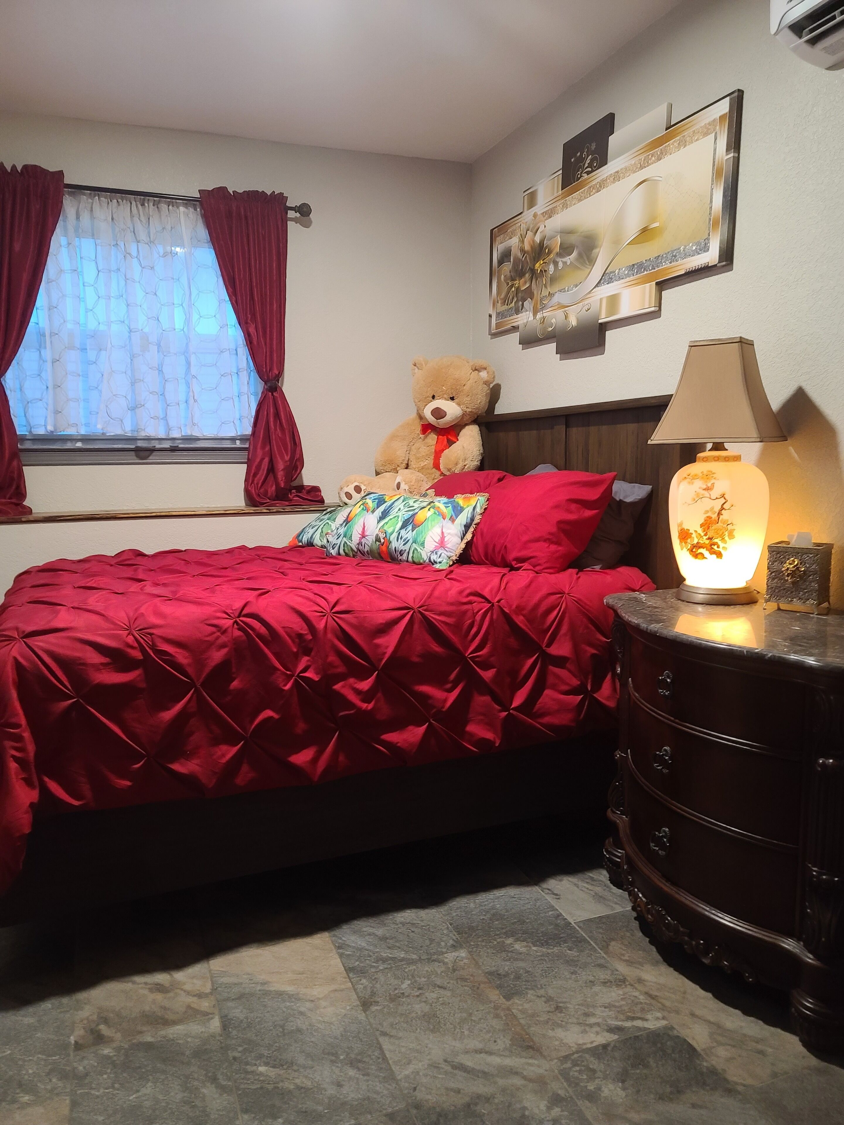 Un dormitorio con una cama, un osito de peluche, una lámpara y un cuadro en la pared.