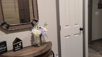 Un pasillo con una puerta blanca, un espejo con marco de madera, un tocador curvo con un florero y un cartel de bienvenida.