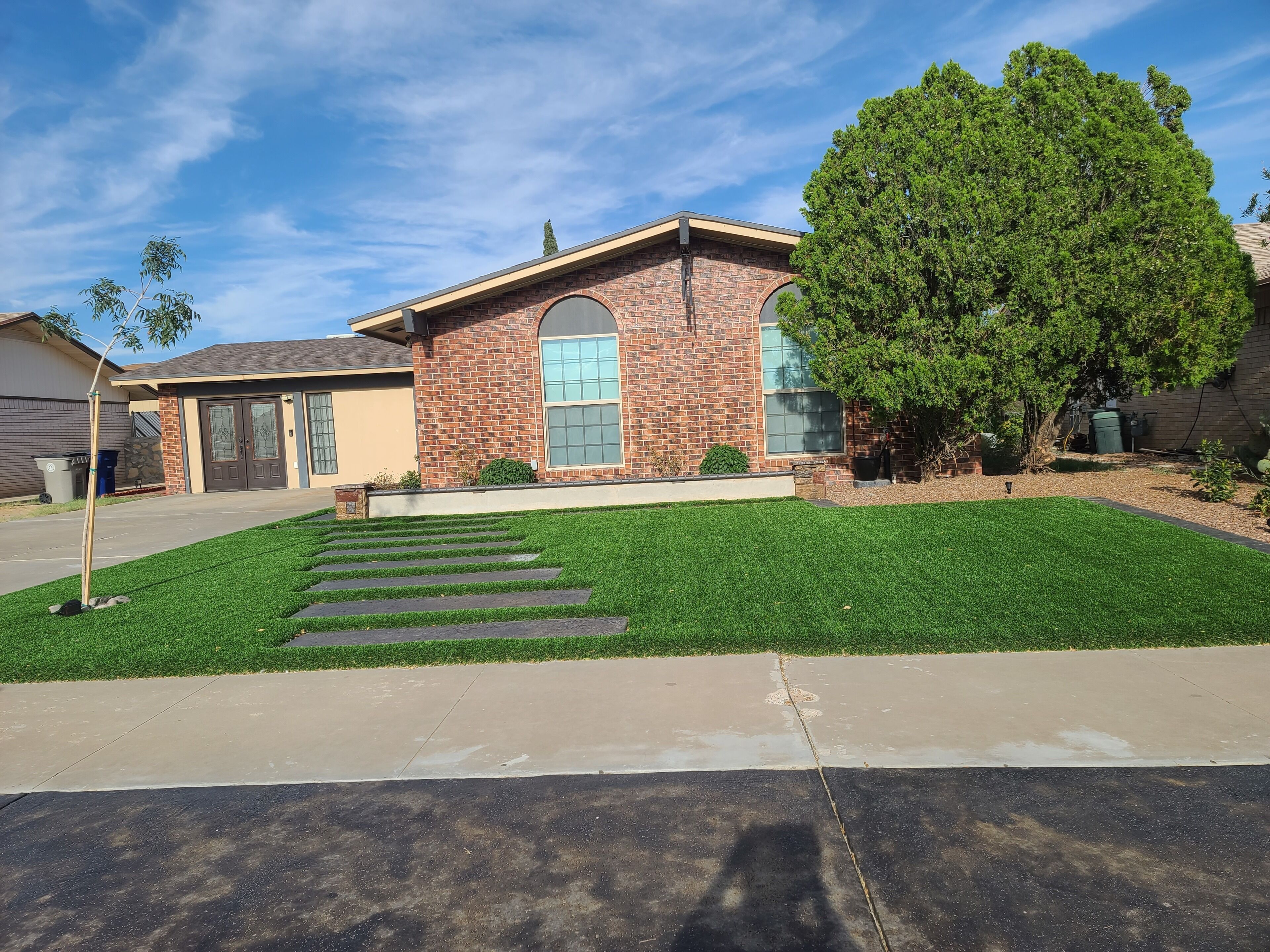 A residential house with a brick facade, a front lawn with a stone pathway, and a large tree to the right.