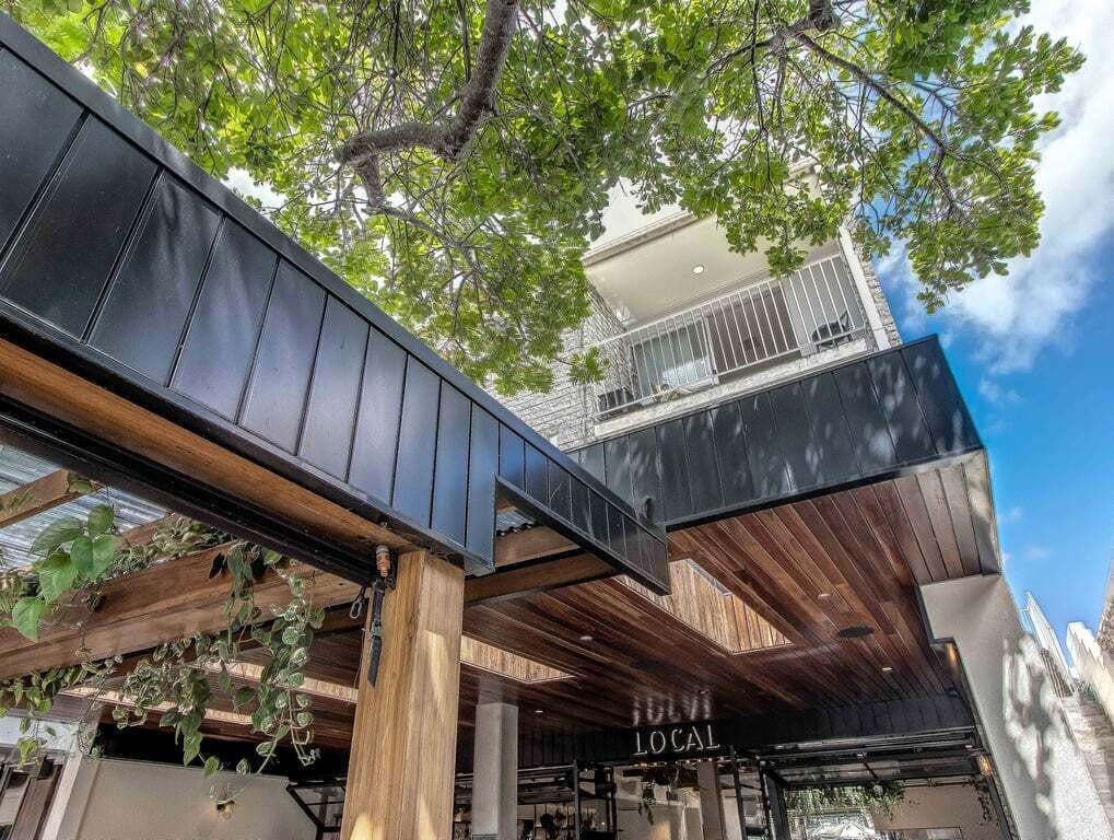 An outdoor area with a wooden pergola, a sign reading 'LOCAL', and a building with a balcony.