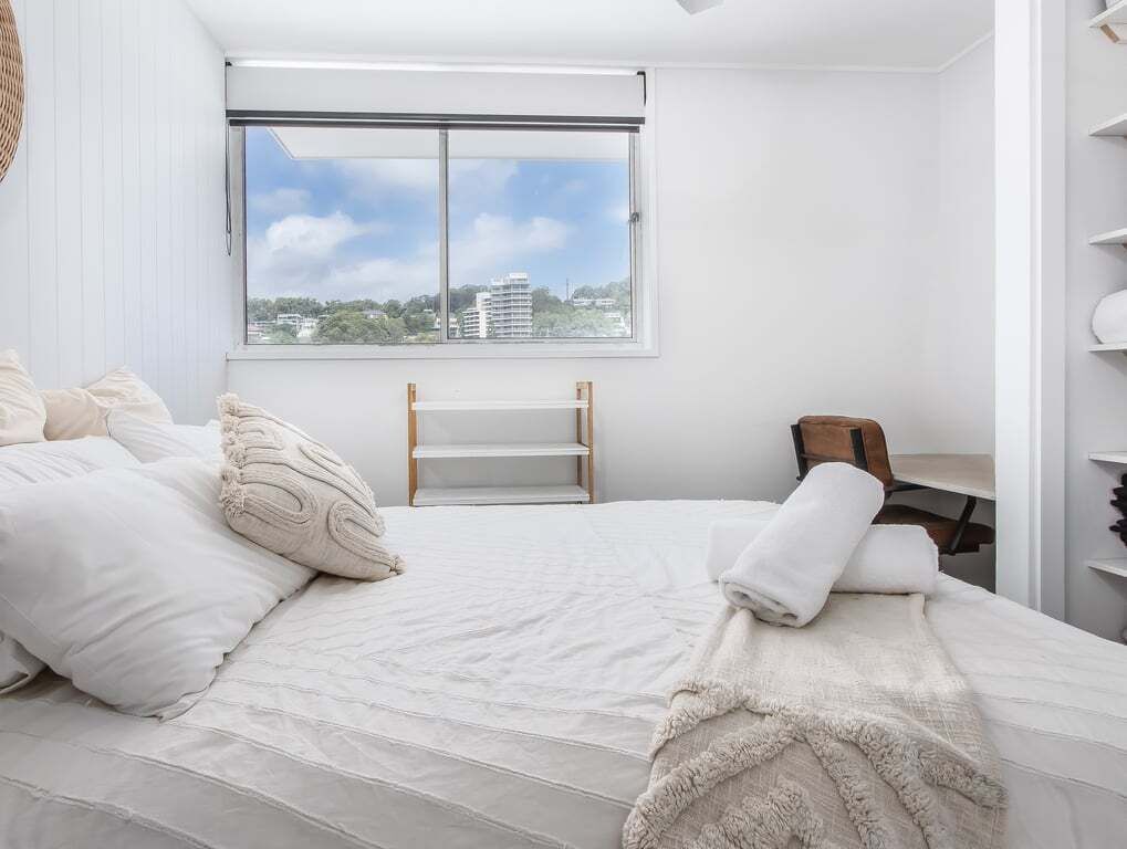 Un dormitorio con un ventanal grande, una cama con ropa de cama blanca y un banco de madera.