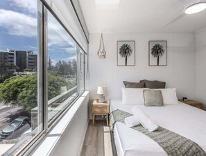 A modern hotel room with a large window, a bed with white linens, a bedside table with a lamp, and two framed pictures on the wall. - The Local Burleigh Heads (Gold Coast)